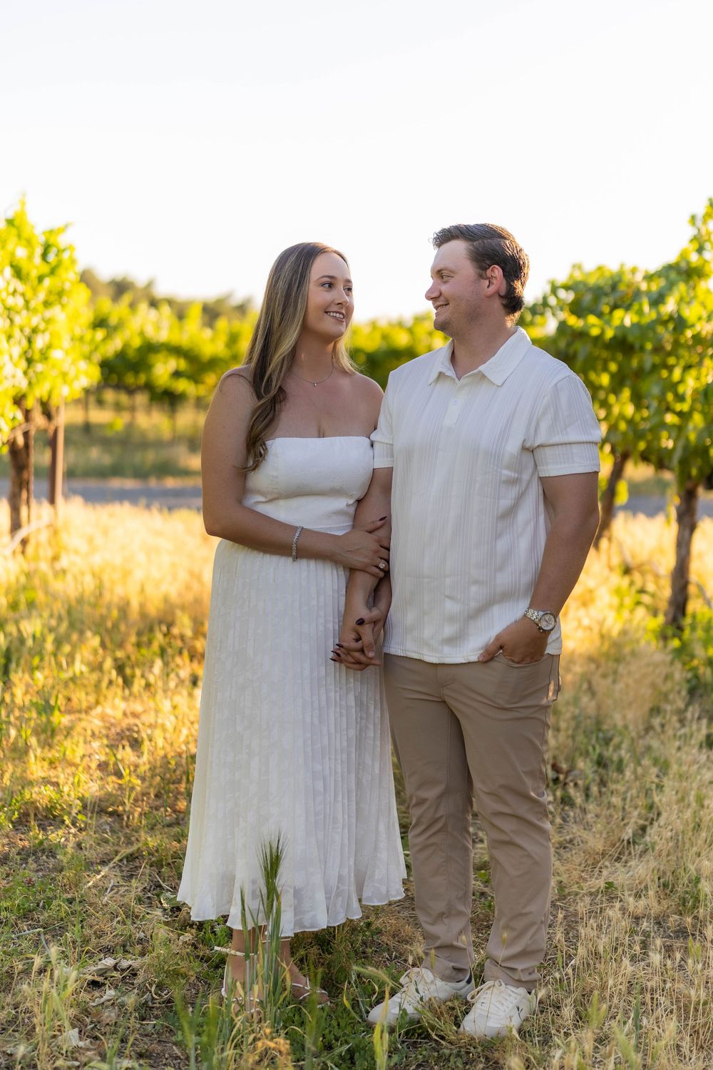 Happy couple in white outfits holding hands in a sunlit vineyard at sunset.