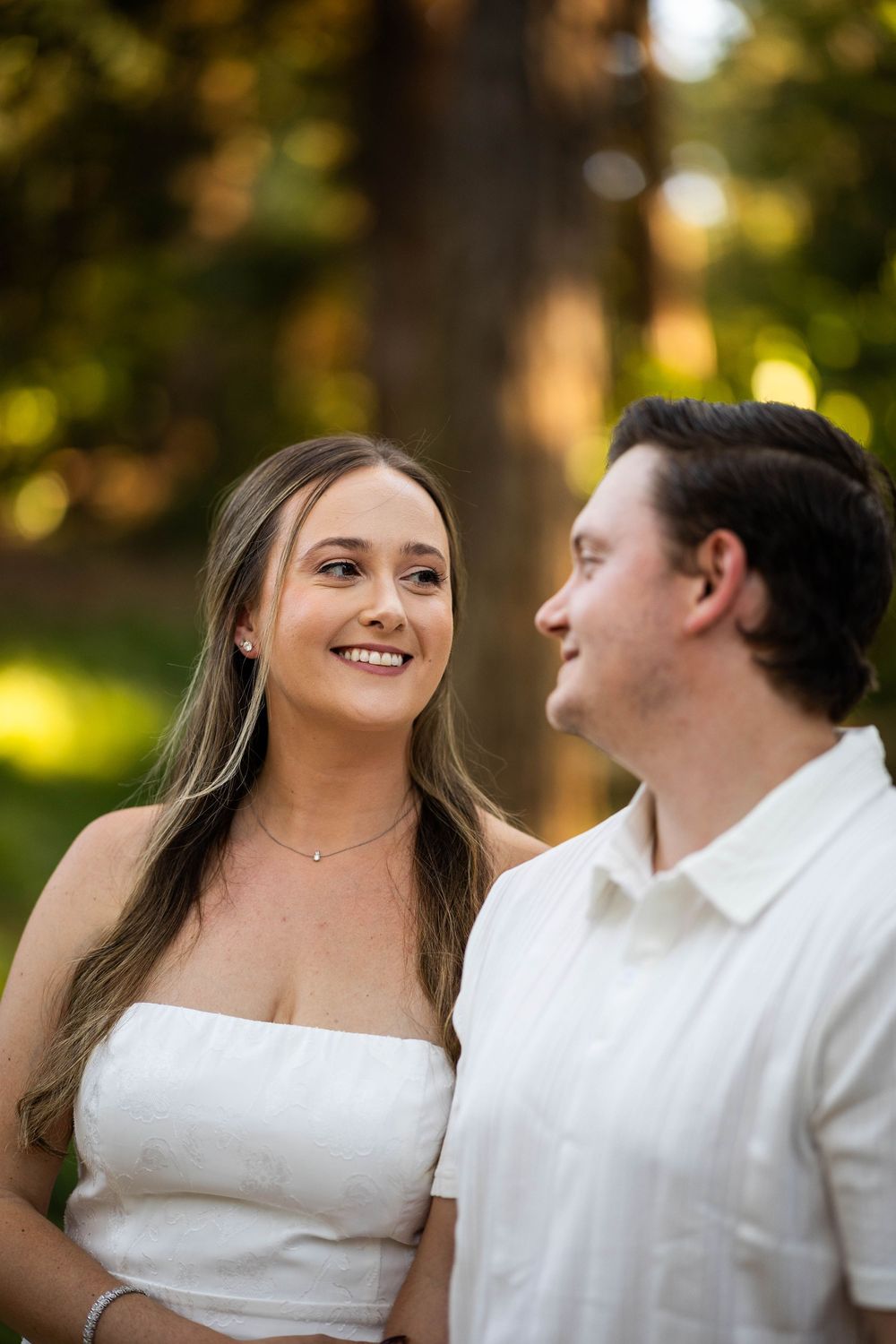 A smiling couple in white outfits share a romantic moment outdoors, surrounded by lush green trees in soft golden light.