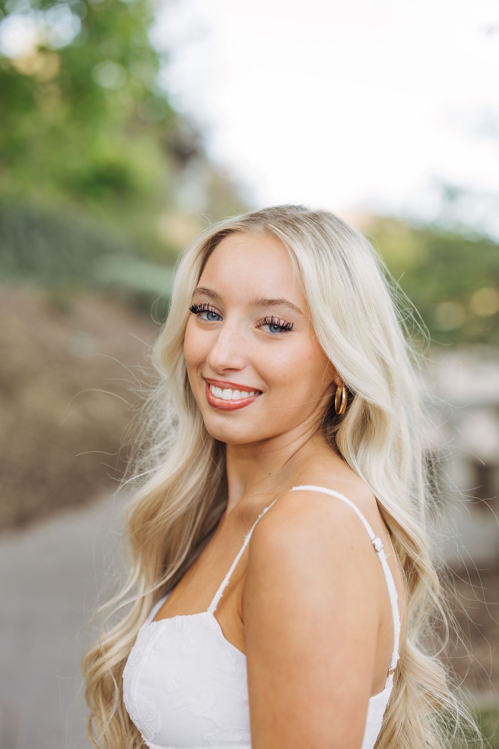 Beautiful blonde young woman smiling outdoors in a white dress with lush greenery behind her.