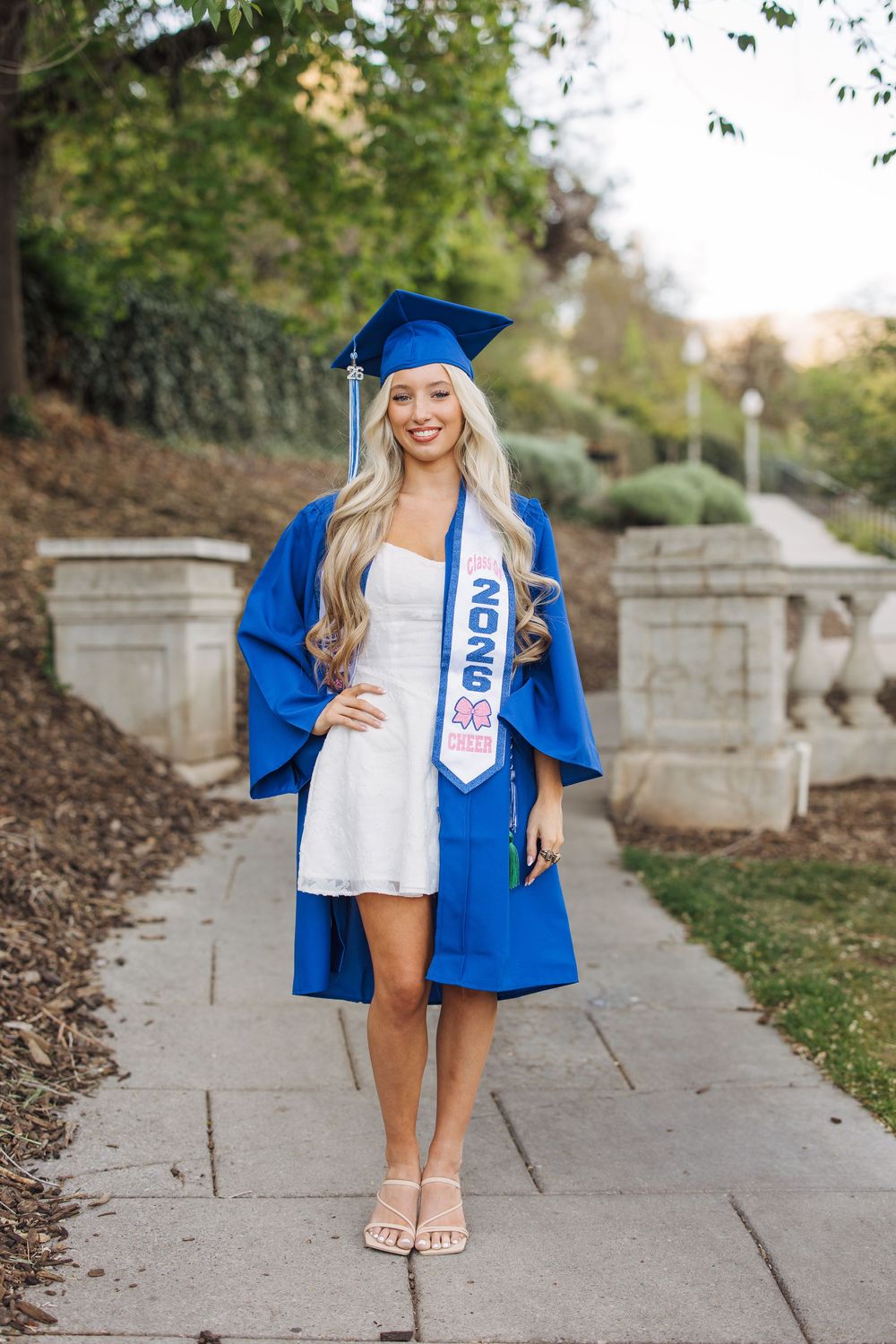 Blonde graduate in blue cap and gown with 2025 stole posing on a tree-lined campus walkway.