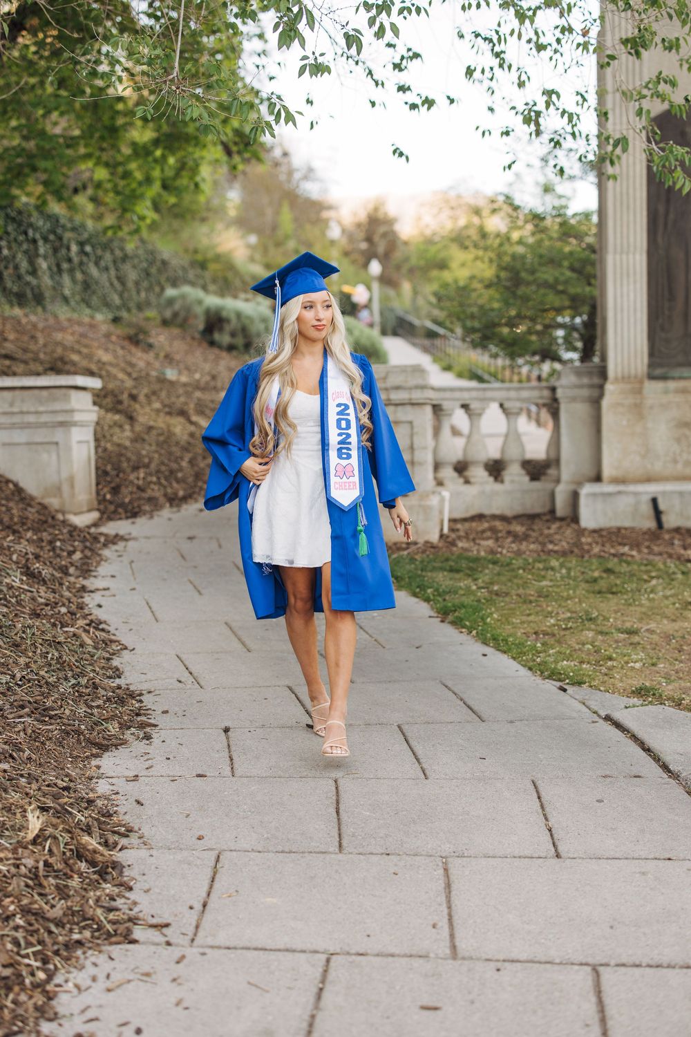Blonde graduate in blue cap and gown walking confidently along a stone campus pathway.