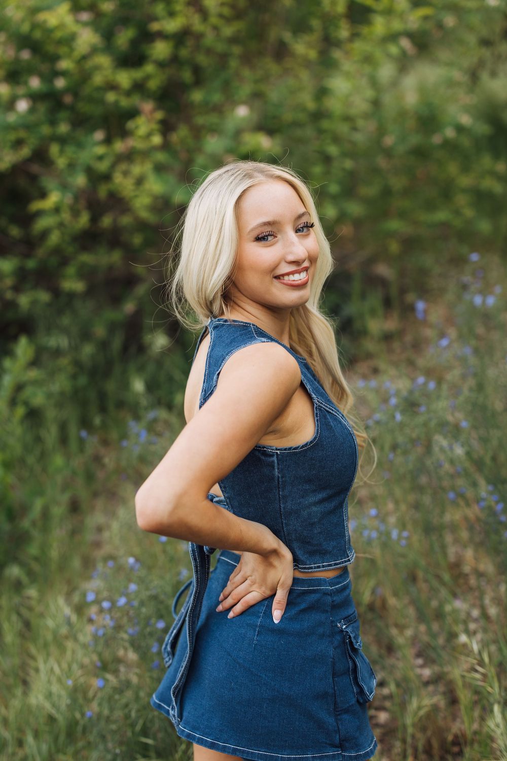 Smiling blonde woman in denim crop top and mini skirt posing outdoors among blue wildflowers and green foliage.
