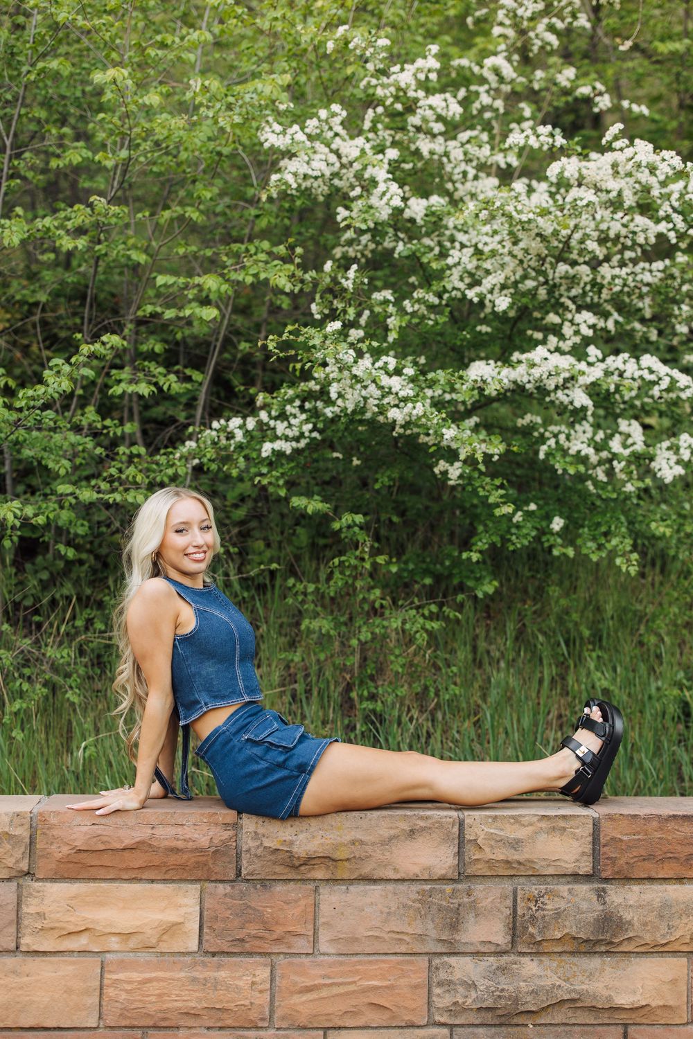 Blonde woman in denim outfit sitting on stone wall with blooming white flowering tree in background.