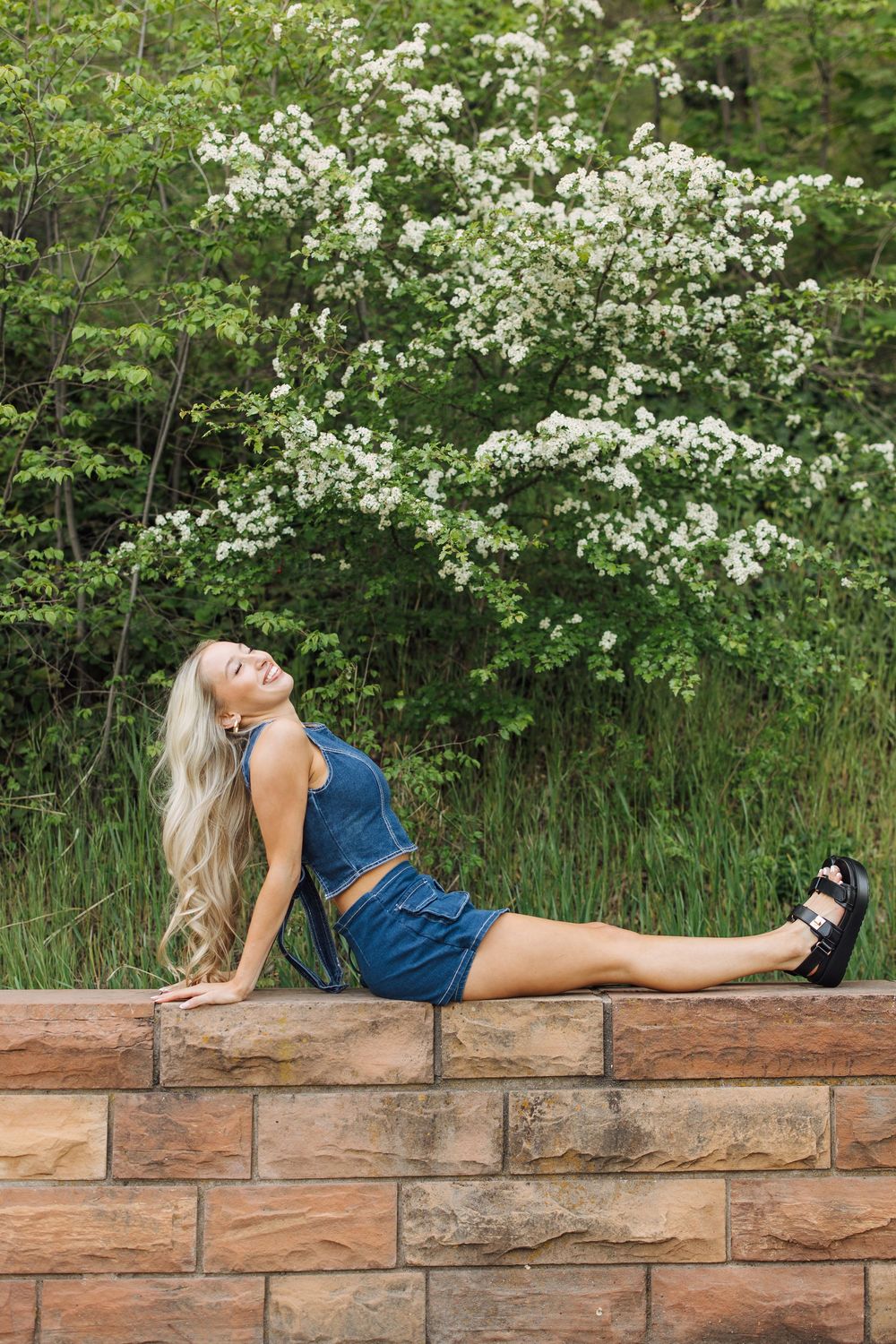 Blonde woman in denim crop top and skirt reclining on stone wall with blooming white trees behind her.