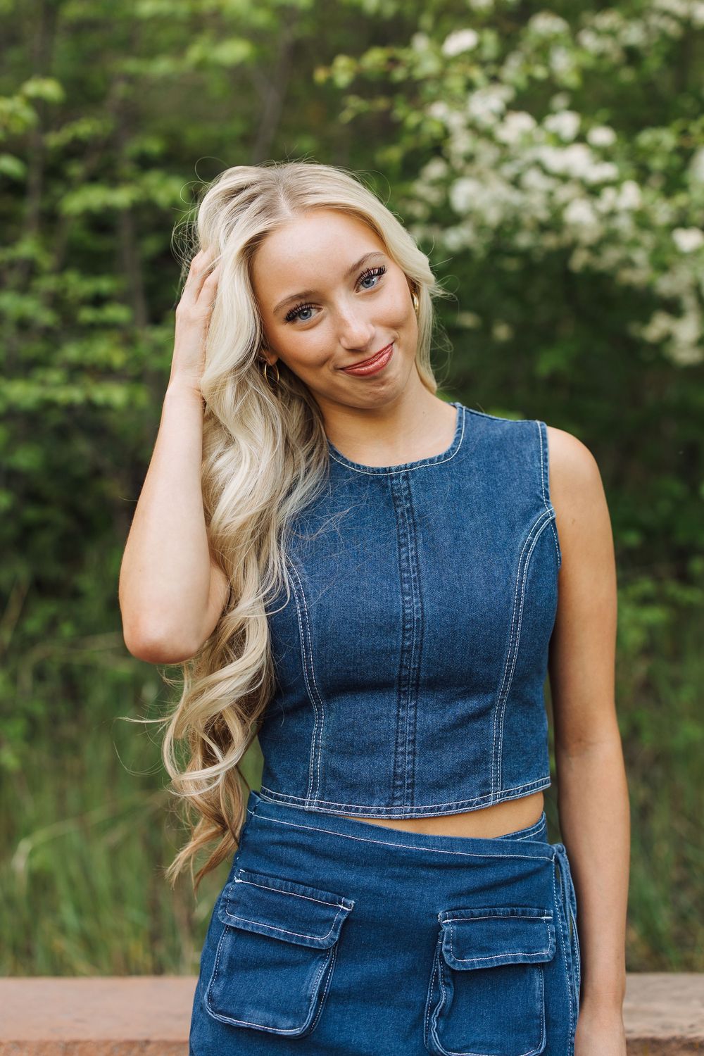 Smiling blonde woman in denim crop top and skirt touching her hair outdoors near white flowering trees.