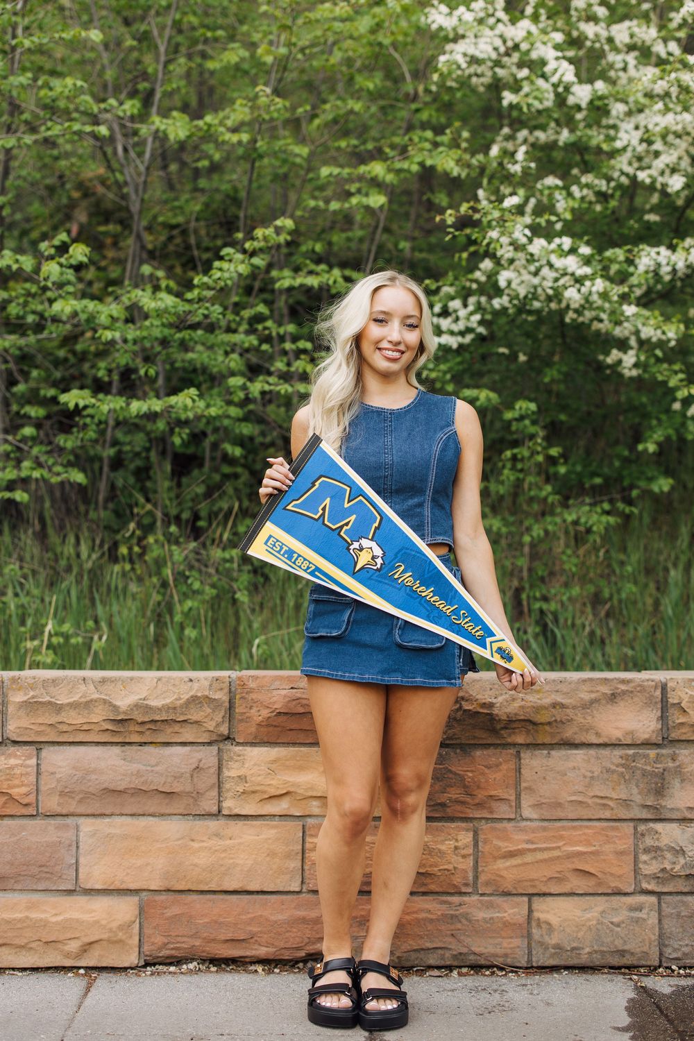 Blonde woman in denim dress holding Morehead State University pennant flag while standing by stone wall outdoors.