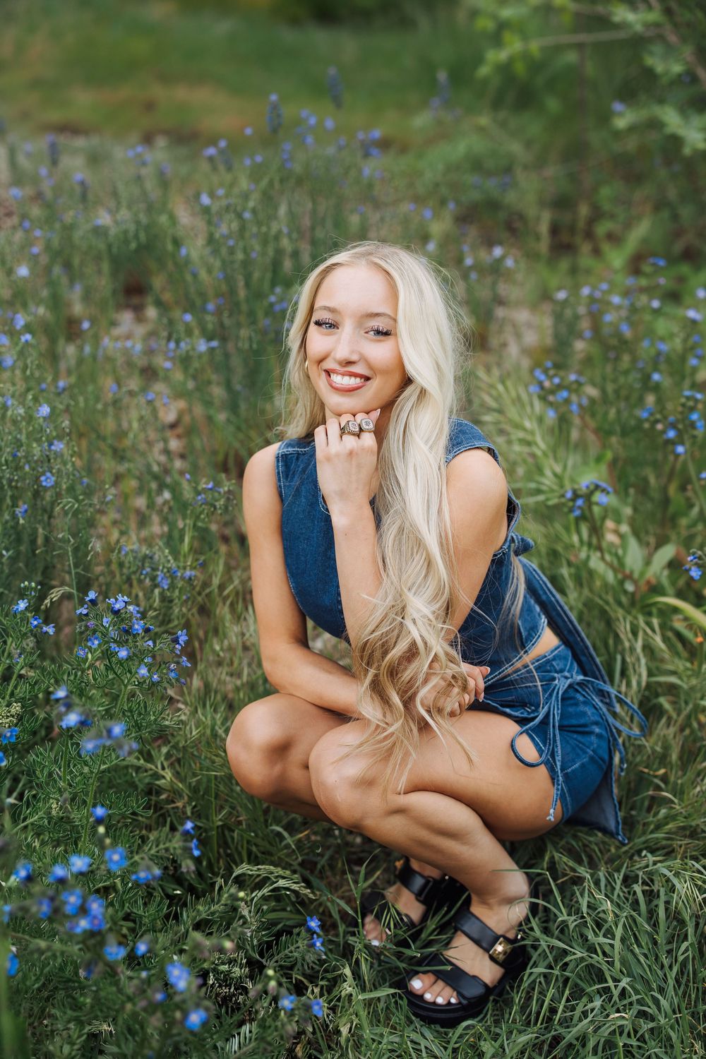 Smiling blonde woman in denim outfit crouching among blue wildflowers in a lush green outdoor setting.
