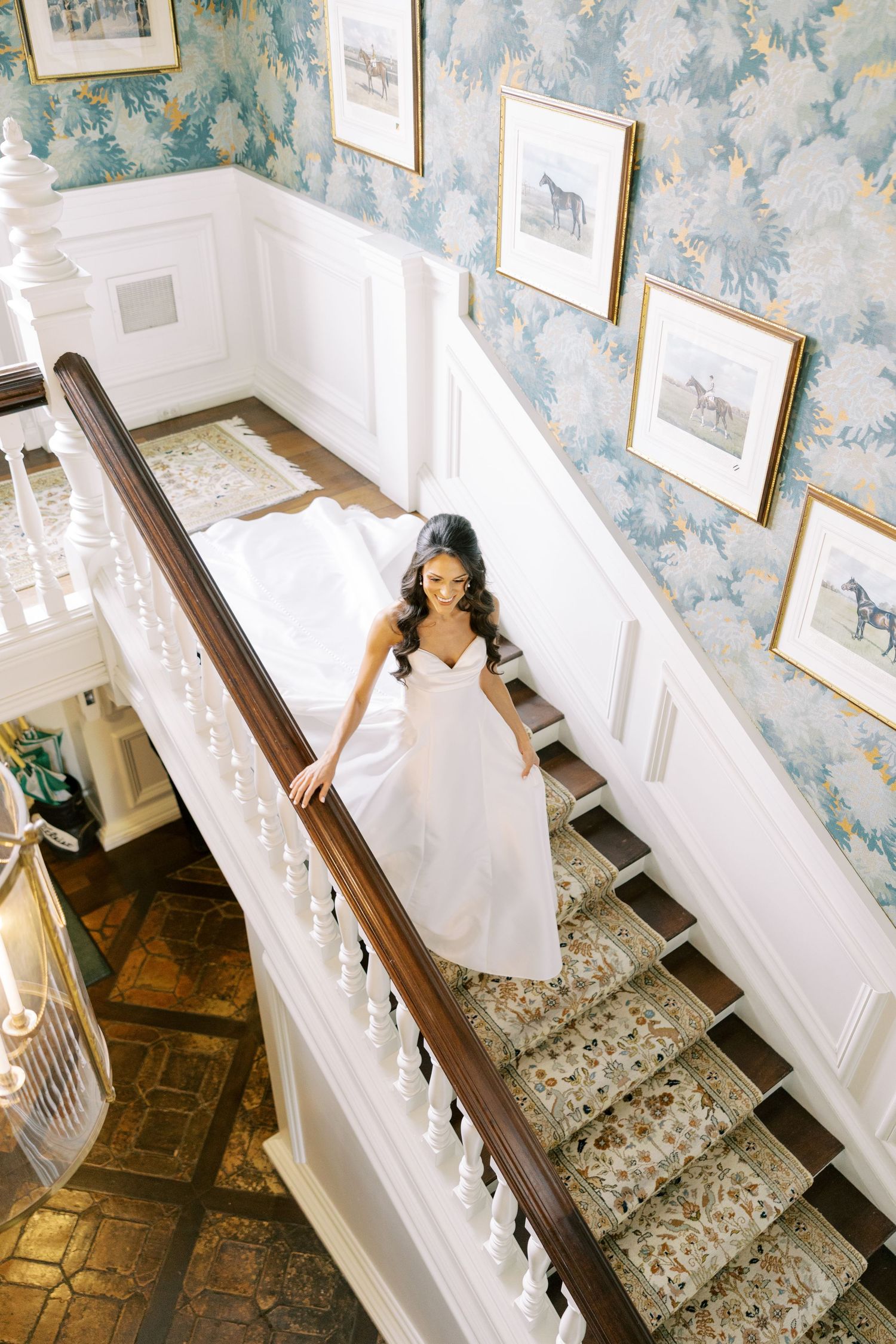 Bride in white gown descends elegant staircase with floral wallpaper and framed art in a classic estate home.