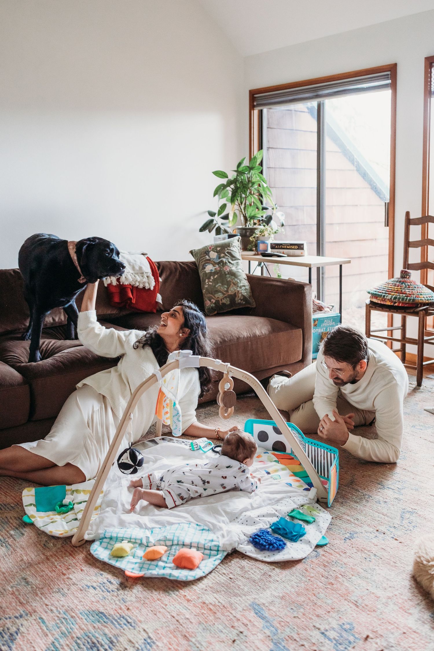 Family with baby on play mat, dog on couch, cozy living room with large windows and warm natural light.