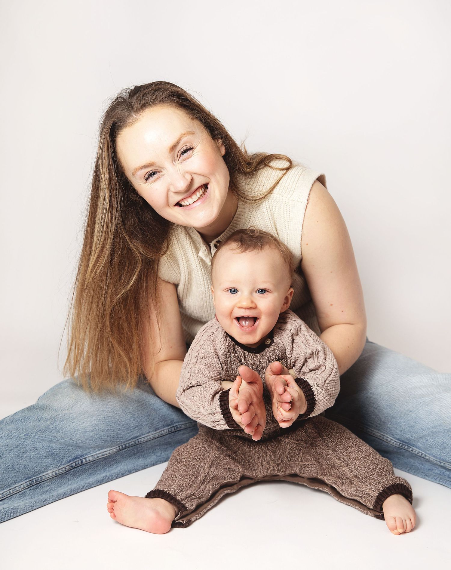 Smiling woman sitting with happy laughing baby in brown outfit against white background. During a studio baby photography session in Bexley