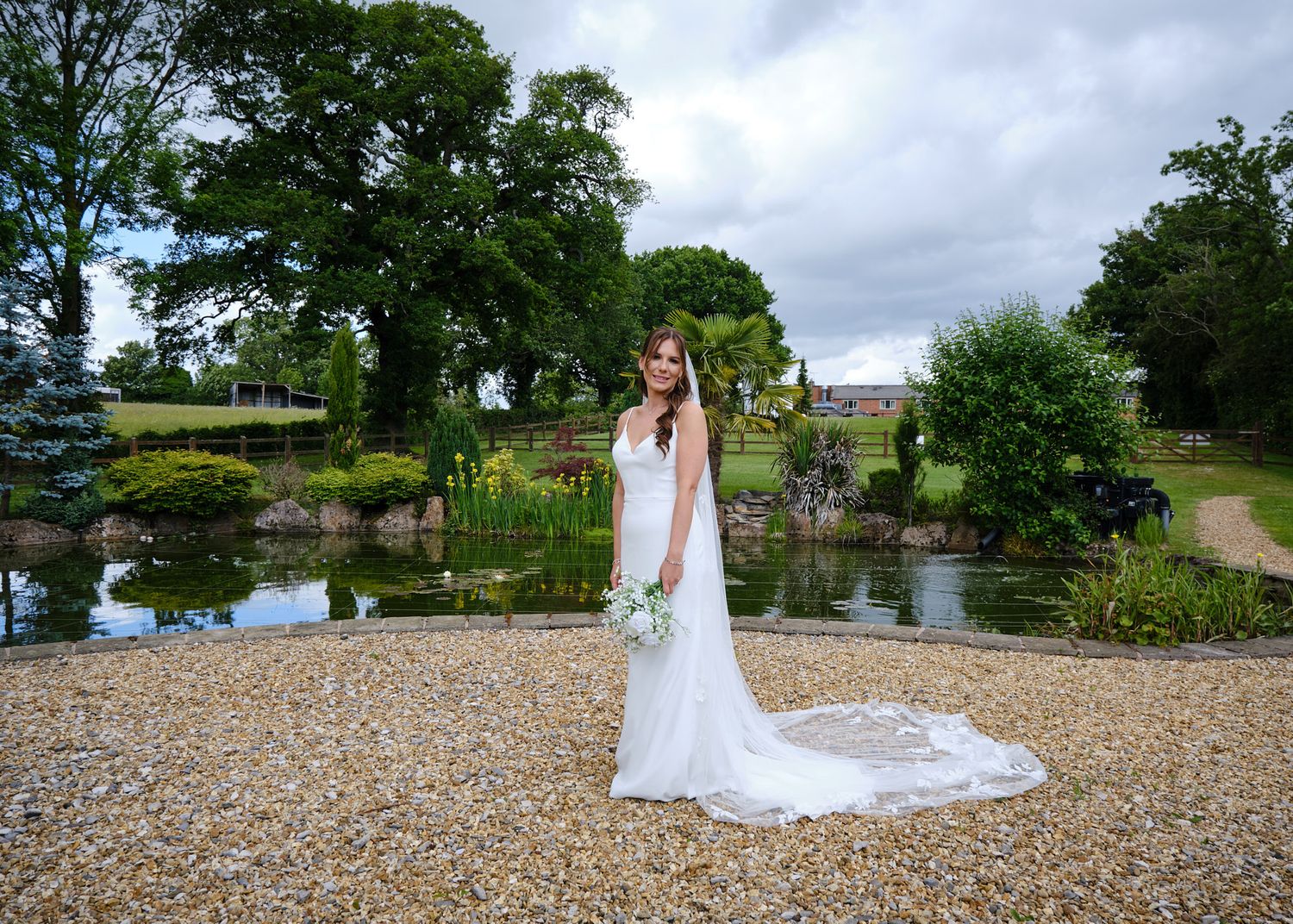 Bride in white gown with veil holding bouquet stands on gravel path beside a serene garden pond.
