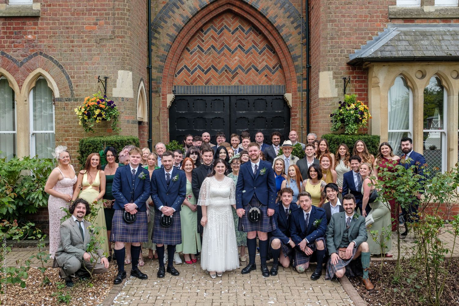 Large wedding group photo outside a brick church, guests in kilts and formal attire with bride and groom centered.