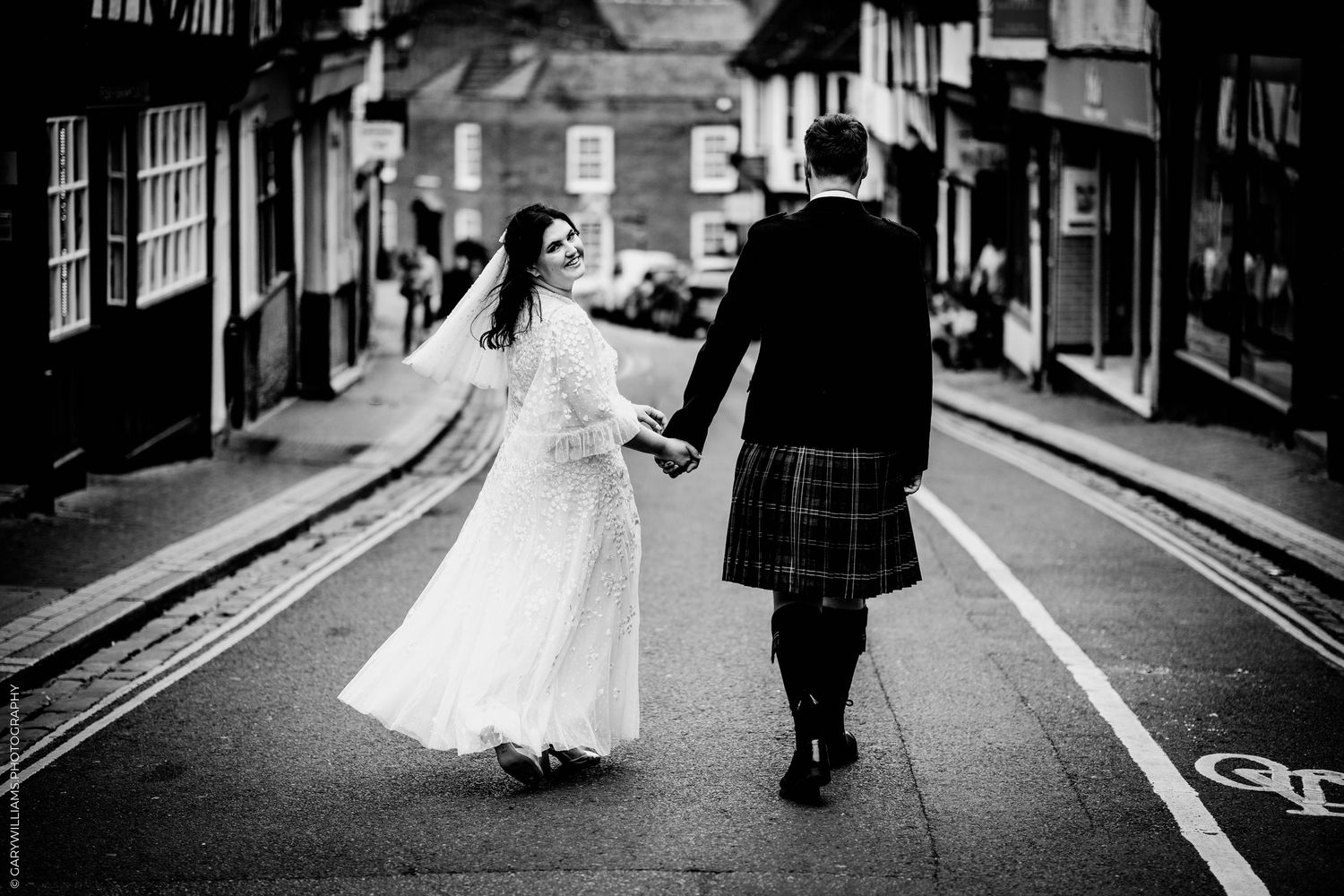 Black and white photo of a bride and groom walking hand in hand down a cobblestone street, groom wearing a kilt.