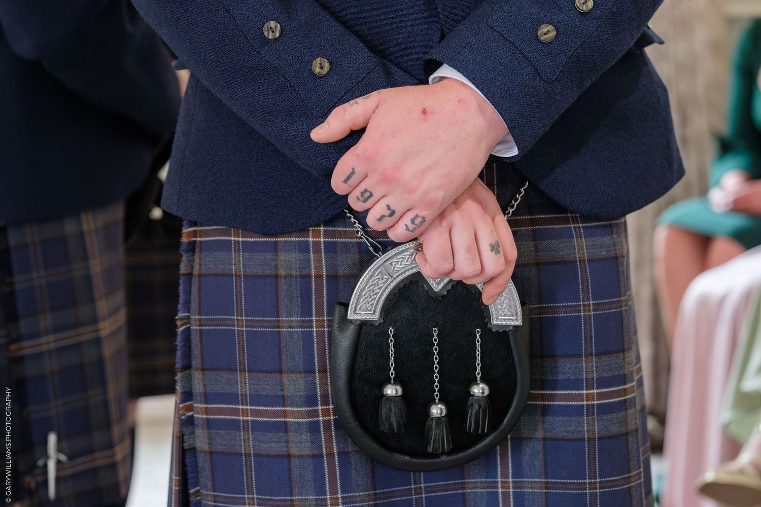 Man in Scottish tartan kilt wearing a traditional black leather sporran with silver chain tassels at a formal event.
