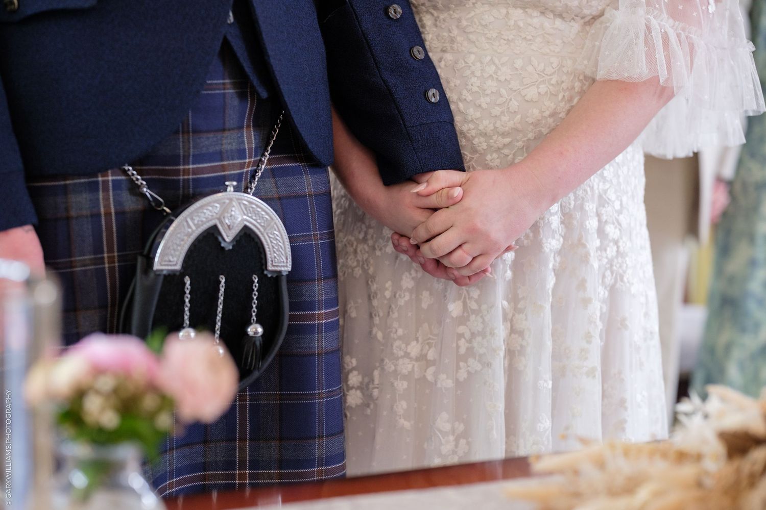 Scottish wedding couple in traditional tartan kilt and lace bridal gown holding hands at ceremony altar.