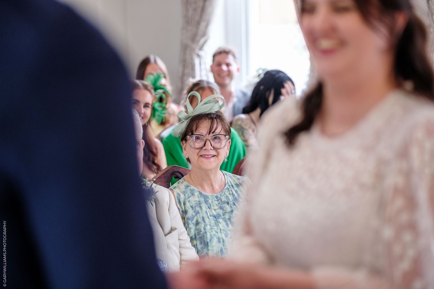 Wedding guests smiling and watching a ceremony in a bright room, woman in green dress and glasses in focus.