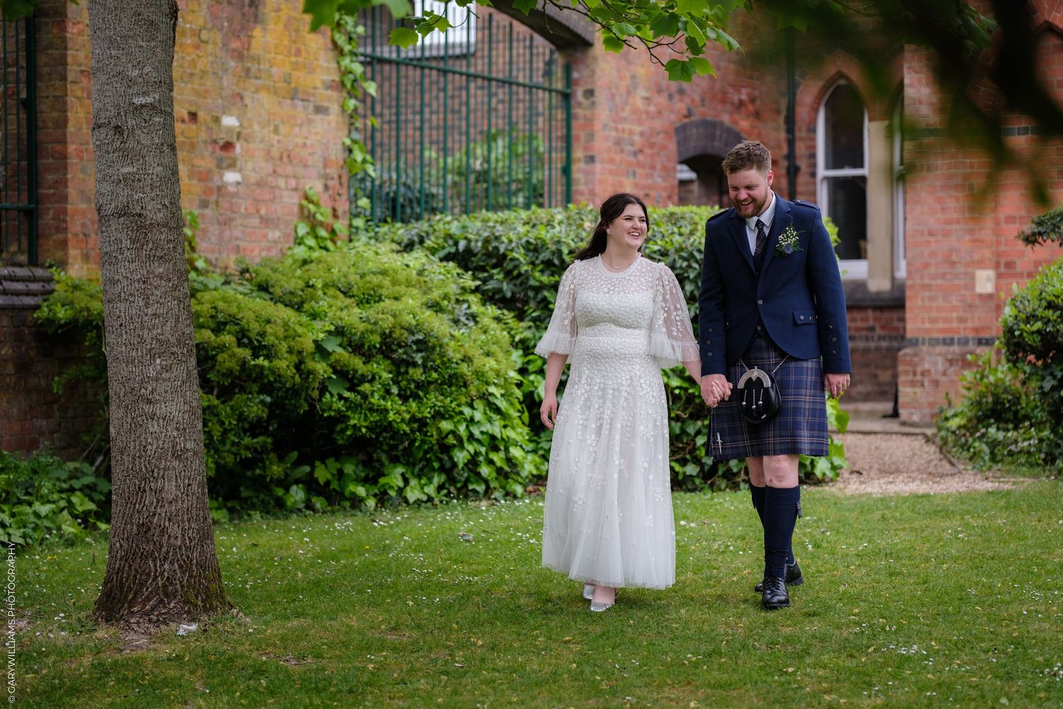 Bride in white lace dress and groom in blue tartan kilt walk hand in hand through a garden.