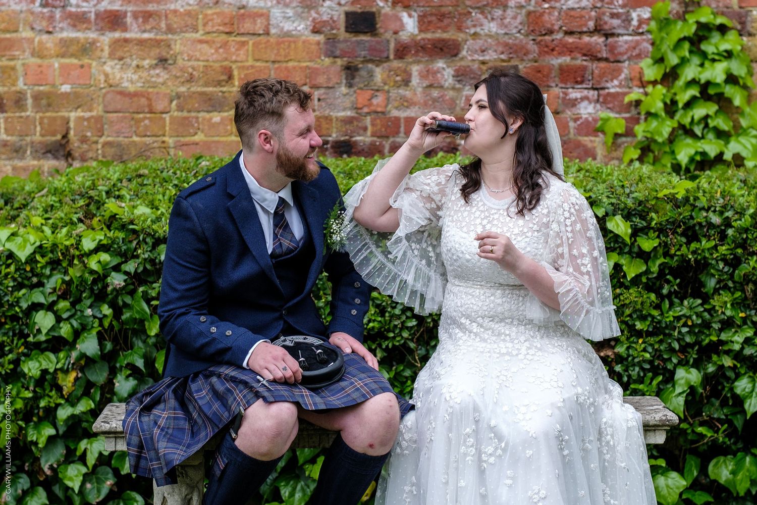 Bride in white lace gown and groom in blue suit with tartan kilt share a drink outdoors at their wedding.