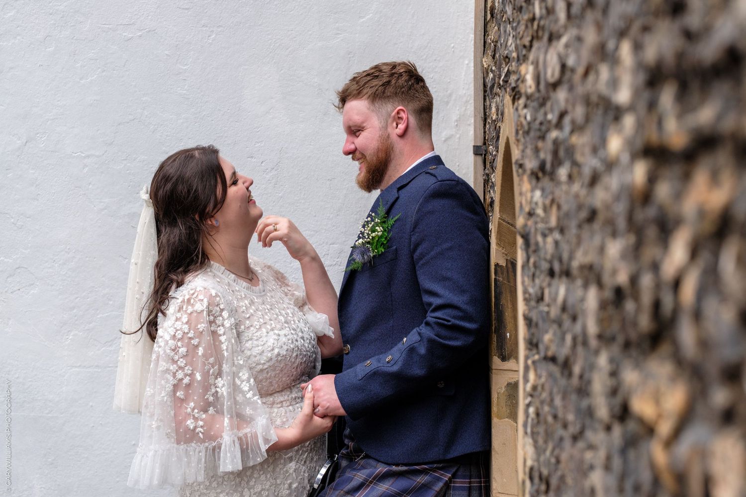 Bride and groom share a tender moment laughing together against a stone wall on their wedding day.