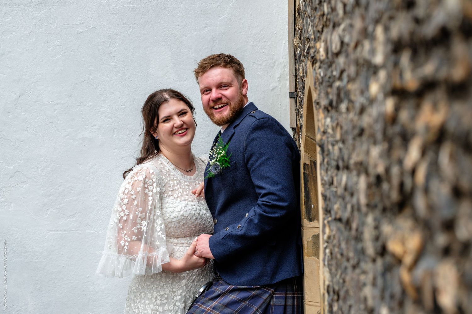 Smiling bride and groom pose together against a white wall, groom wearing a kilt and navy blazer with boutonniere.