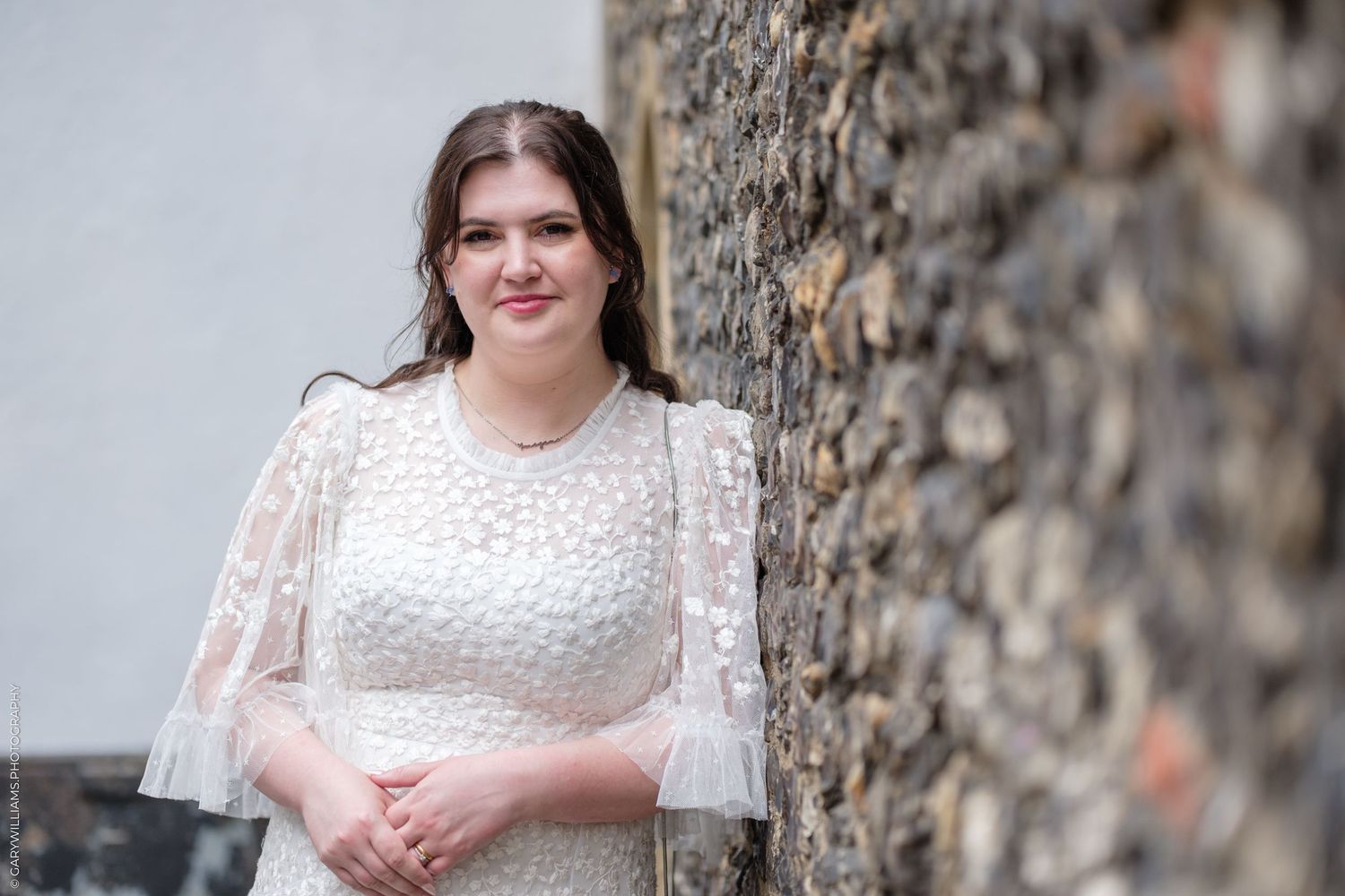 Young woman in white lace dress posing gracefully beside a rustic stone wall outdoors.