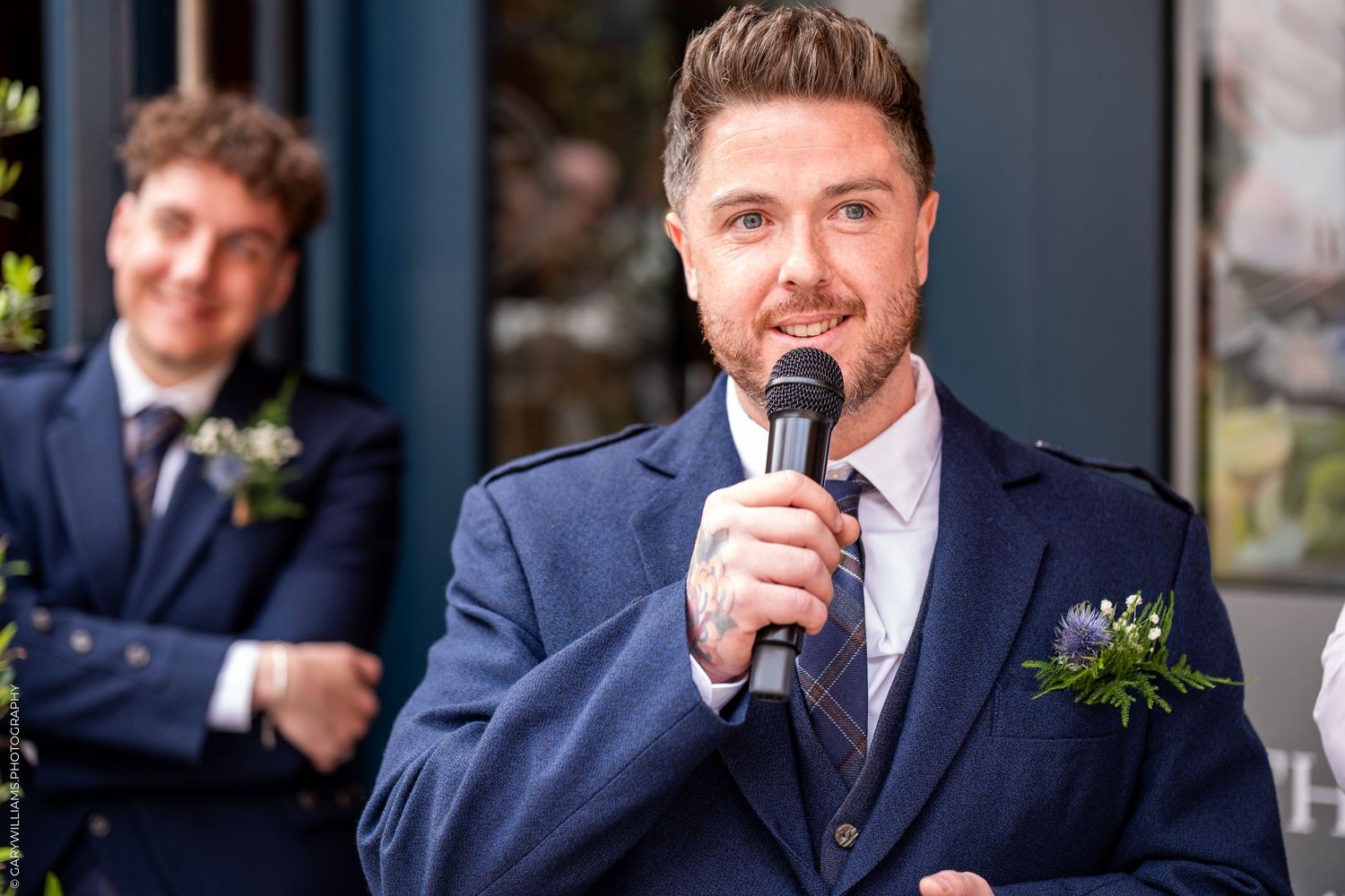 Man in navy suit speaks into microphone at wedding ceremony with boutonniere and guests behind him.