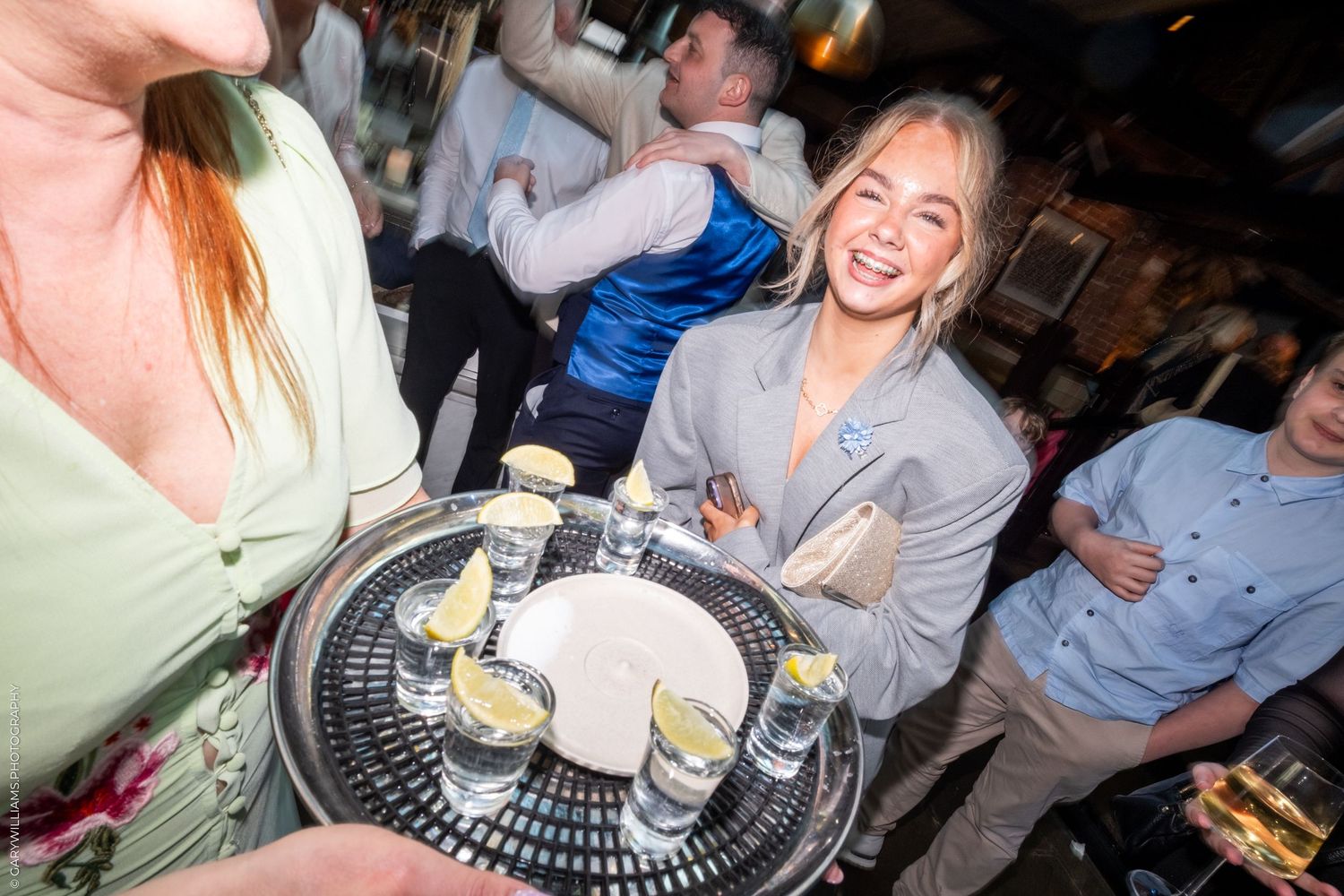 Smiling woman at a bar holding a tray of shot glasses with salt rims at a lively party.
