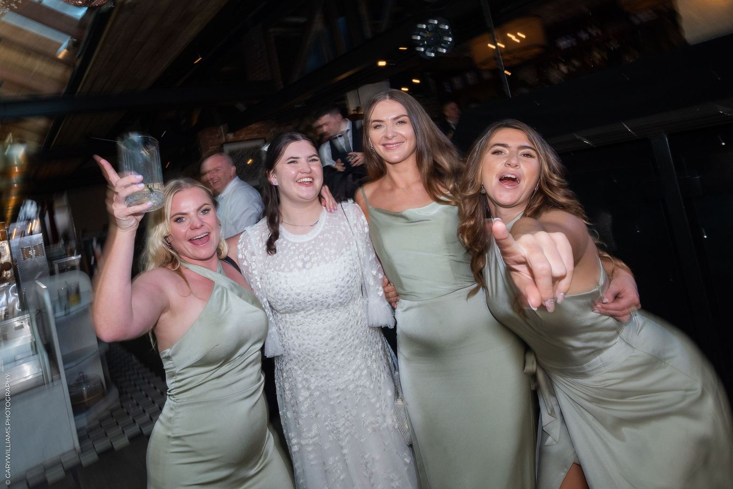 Four women in sage green bridesmaid dresses celebrate with a bride in white at a wedding reception party.