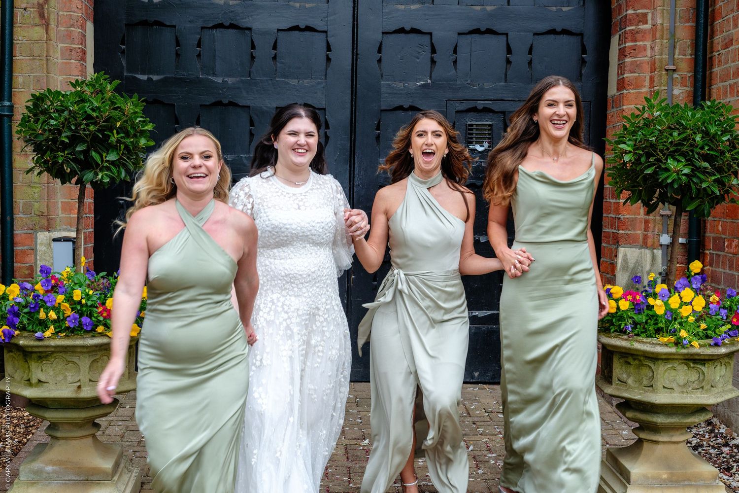 Bride in white sequin gown with three bridesmaids in sage green dresses laughing outside elegant venue doors.