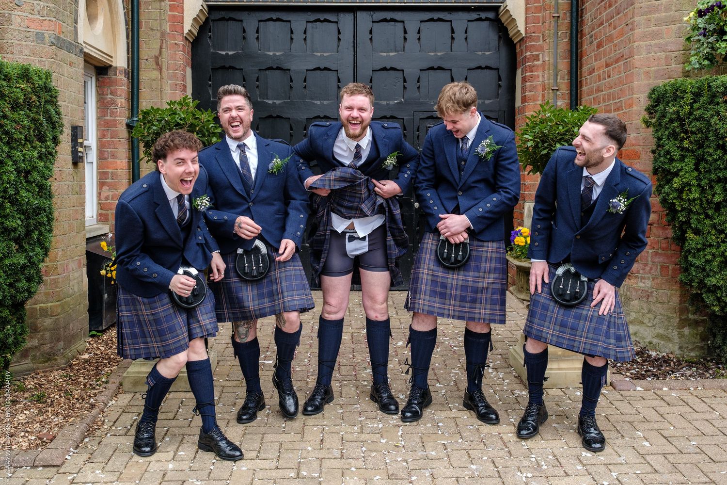 Five men in Scottish kilts and navy blazers laughing together outside a venue door at a wedding celebration.