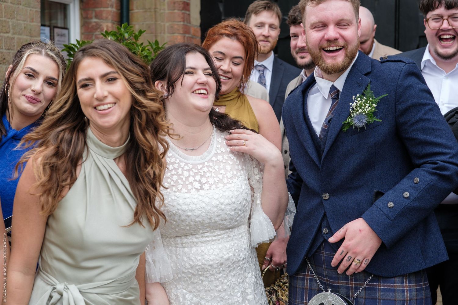 Happy wedding party laughing outside, bride in white dress with groom in Scottish kilt.