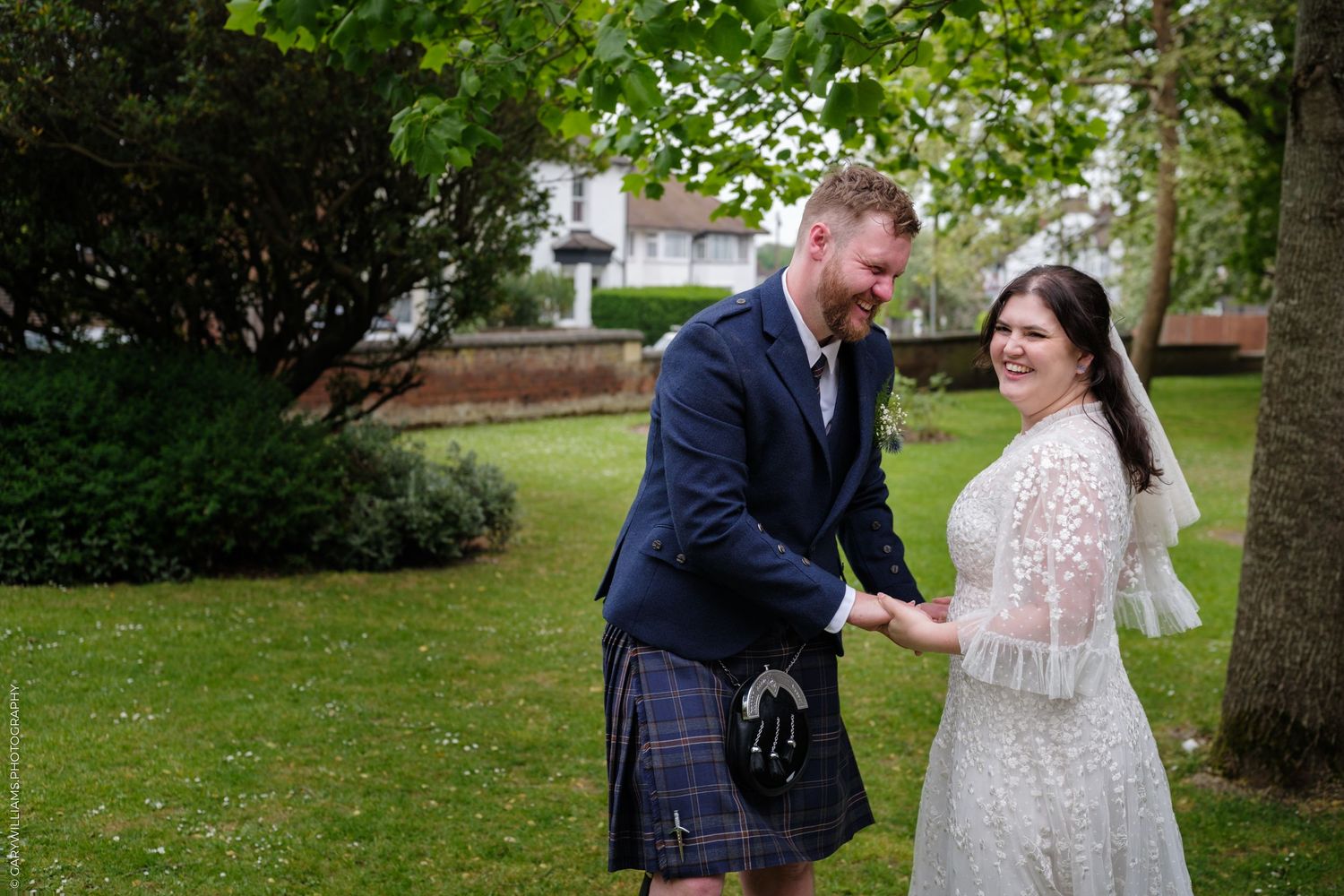 Smiling bride in lace gown and groom in Scottish tartan kilt holding hands outdoors in a lush green garden.