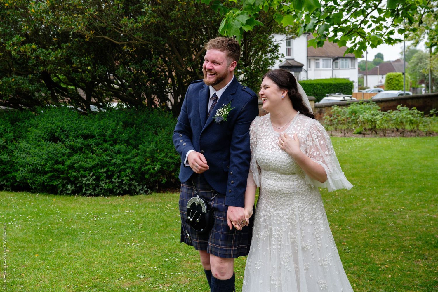 Happy bride and groom in Scottish kilt holding hands, laughing outdoors on their wedding day.
