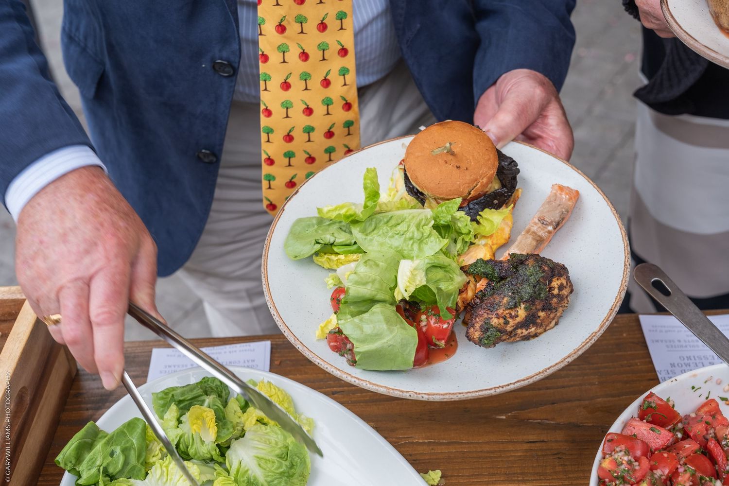 Man in suit serving a plate with burger, fresh greens, and colorful vegetables at an outdoor buffet.