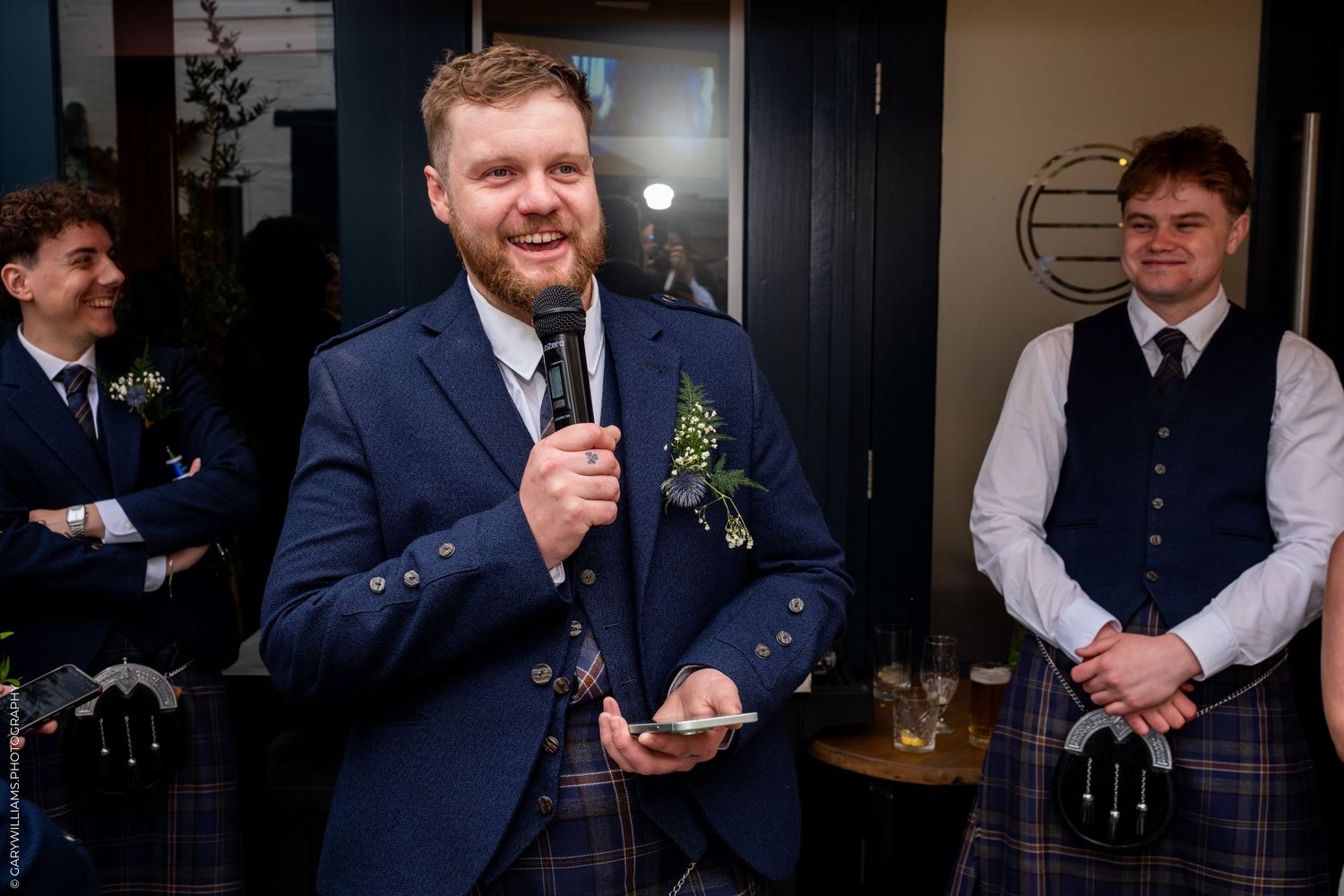 Groom in blue suit and tartan kilt gives a speech while groomsmen smile in the background.