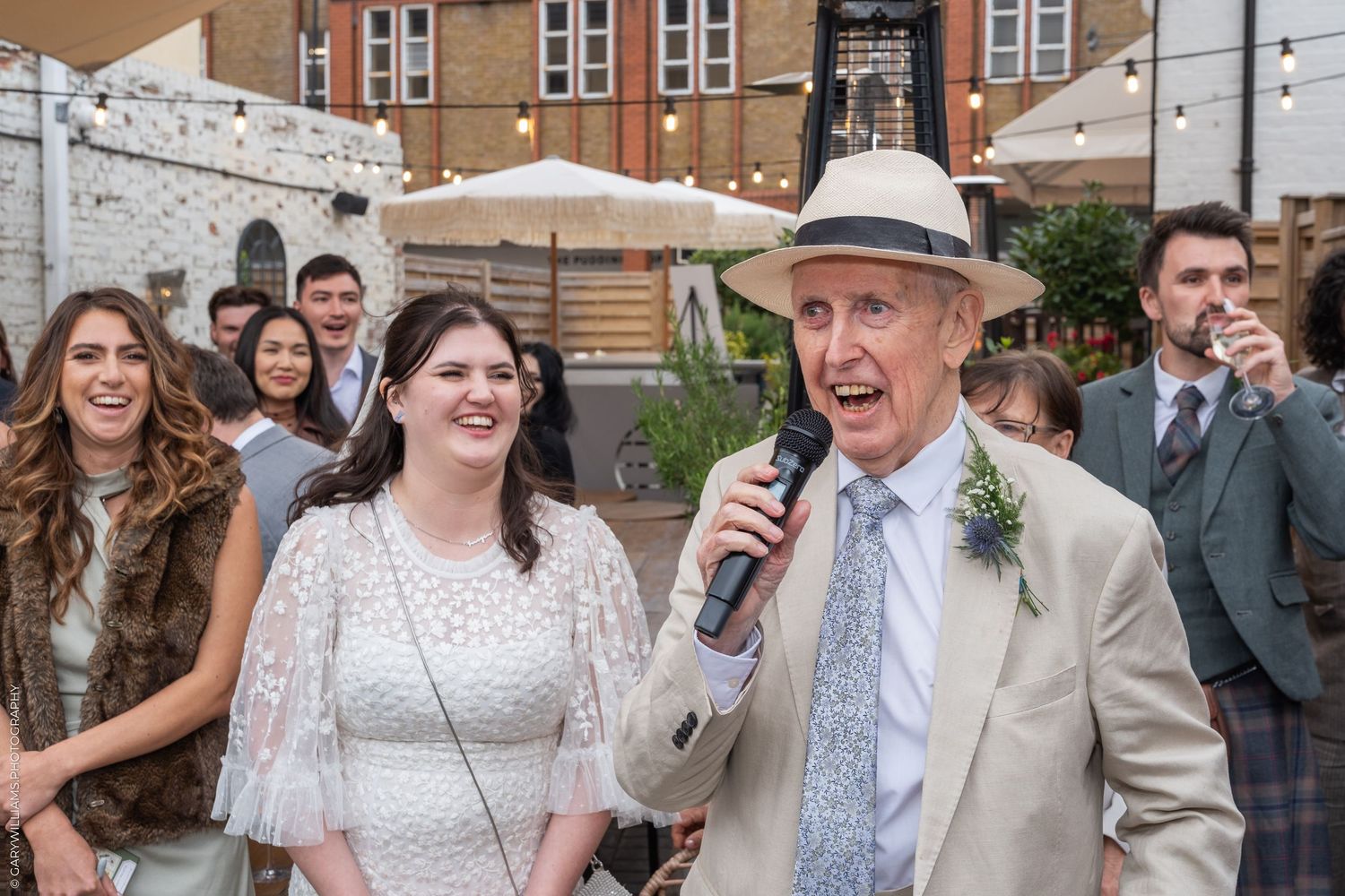 Elderly man in panama hat speaks into microphone at outdoor wedding reception beside smiling bride in white dress.