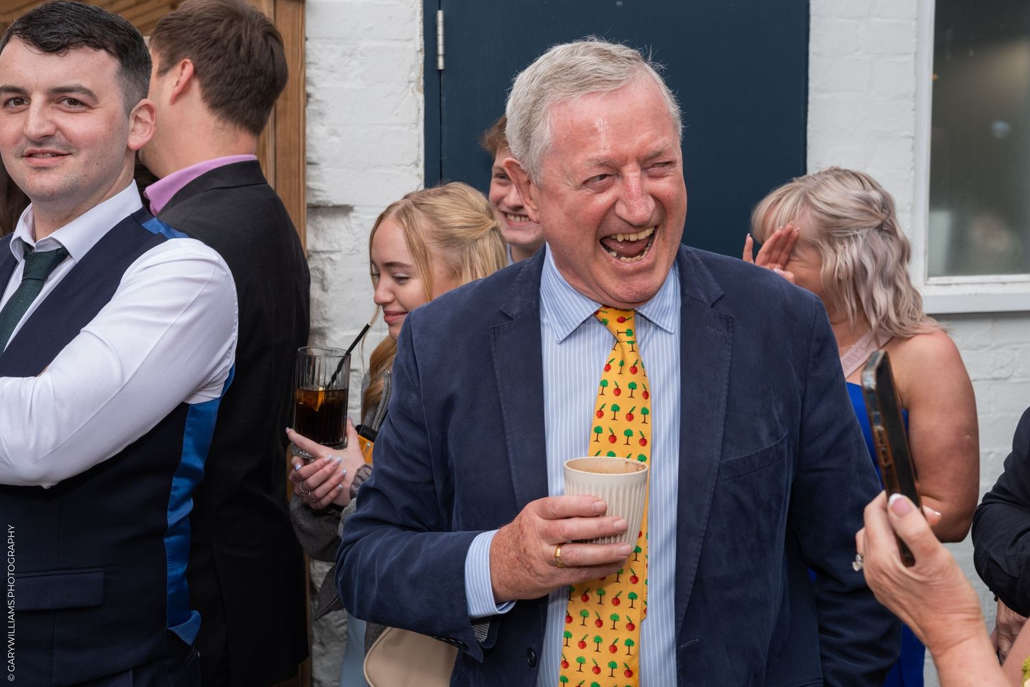Cheerful middle-aged man in blue suit and yellow tie laughing at an outdoor social gathering.