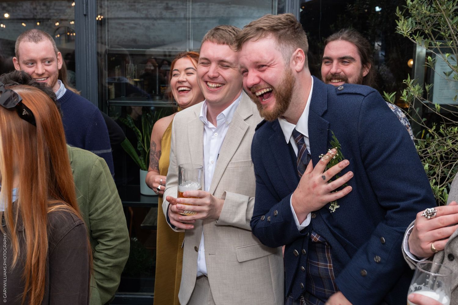 Two men laughing joyfully at a social gathering, one holding a drink, surrounded by smiling guests outdoors.