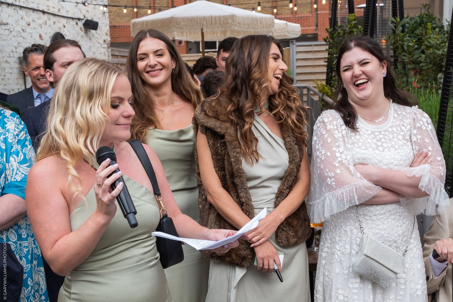 Women in elegant dresses laugh together at an outdoor wedding reception, one holding a microphone and papers.