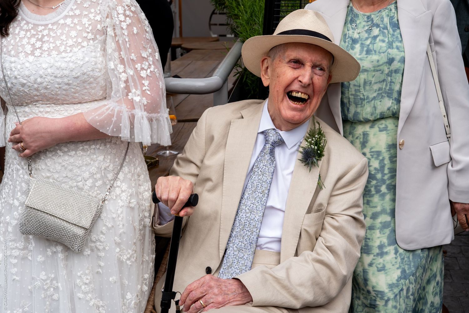 Elderly man in tan suit and hat laughing joyfully in wheelchair at outdoor wedding celebration.