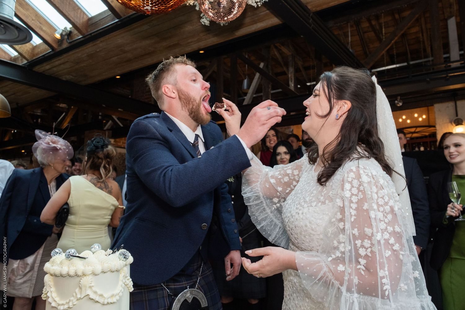 Bride and groom feeding each other wedding cake at their reception in a rustic venue.