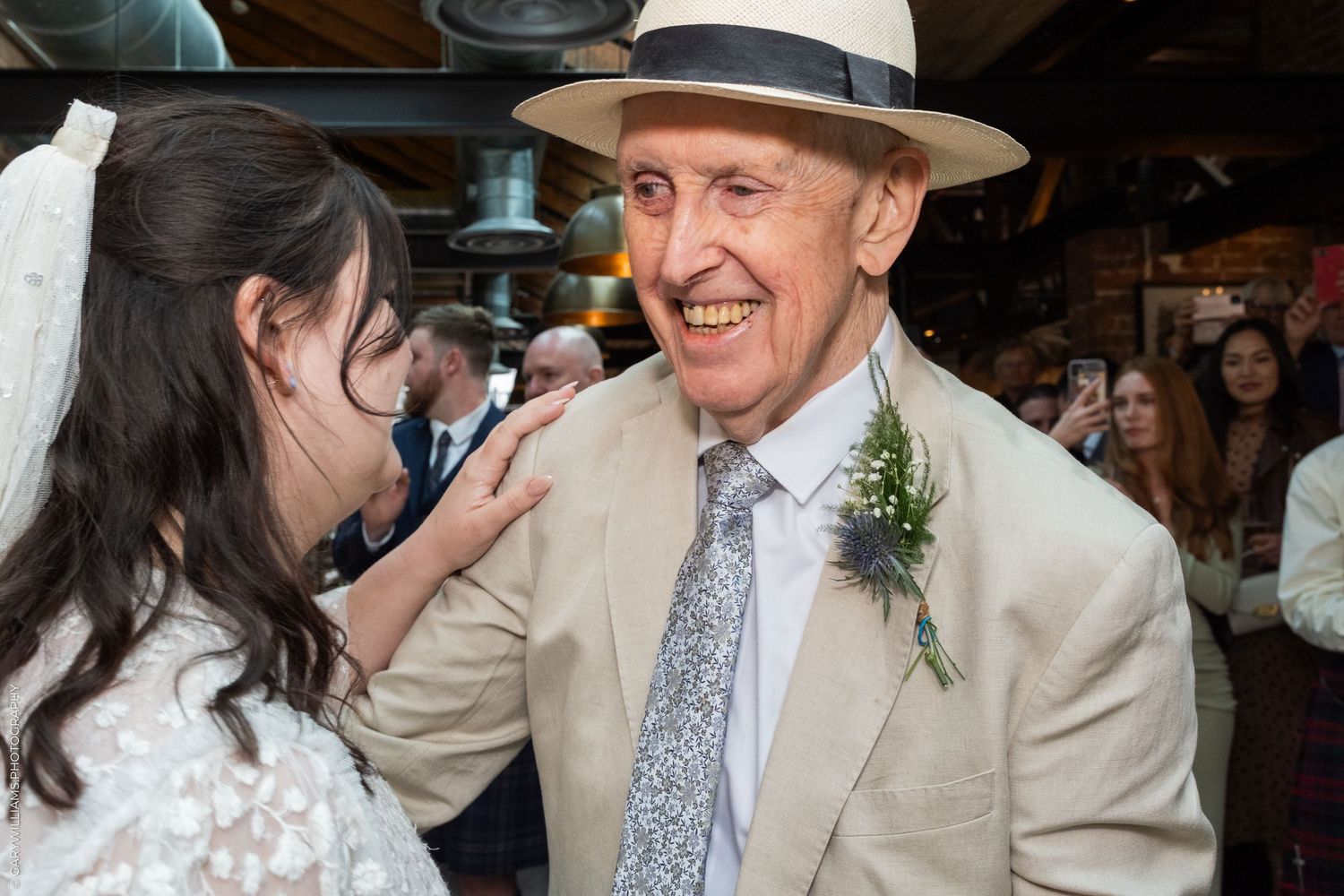 Elderly man in tan suit and hat smiles at bride in white dress at wedding celebration.