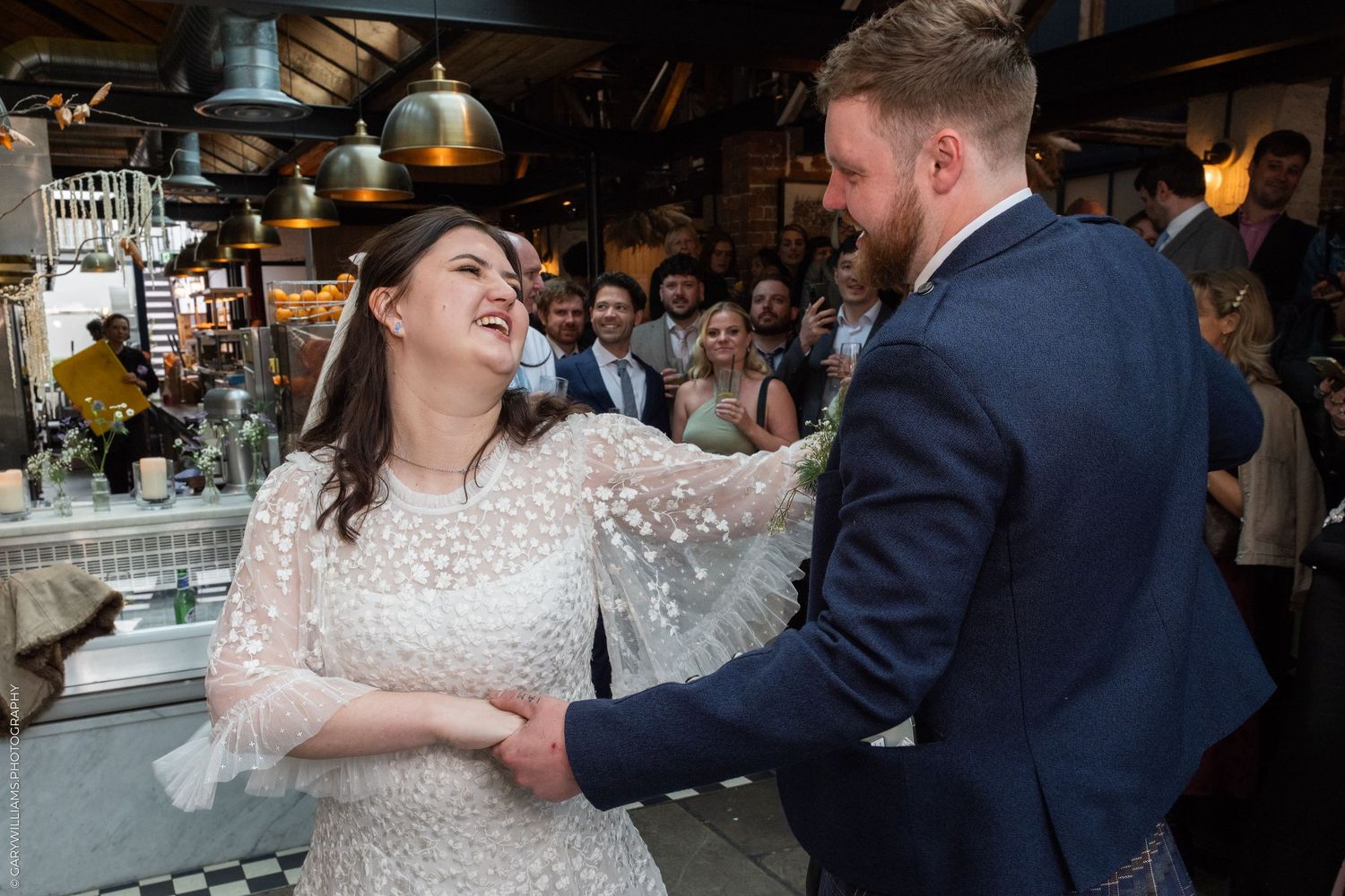 Bride and groom share a joyful first dance at a rustic wedding reception venue filled with guests.