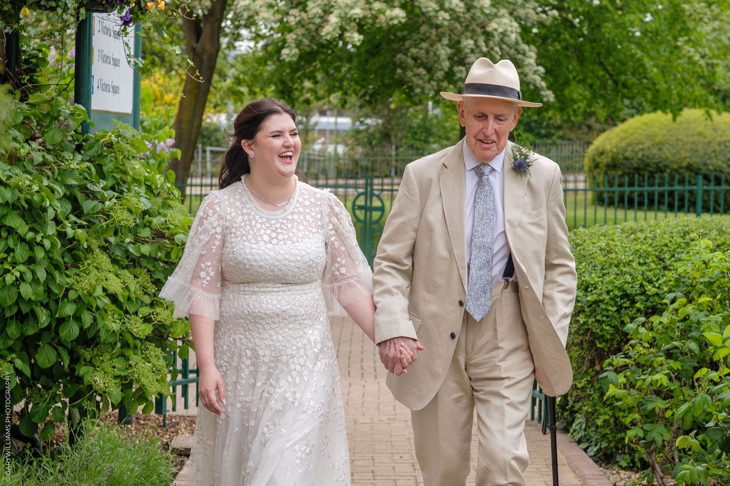Bride in white lace gown walks hand-in-hand with elderly man in tan suit and hat on garden path.