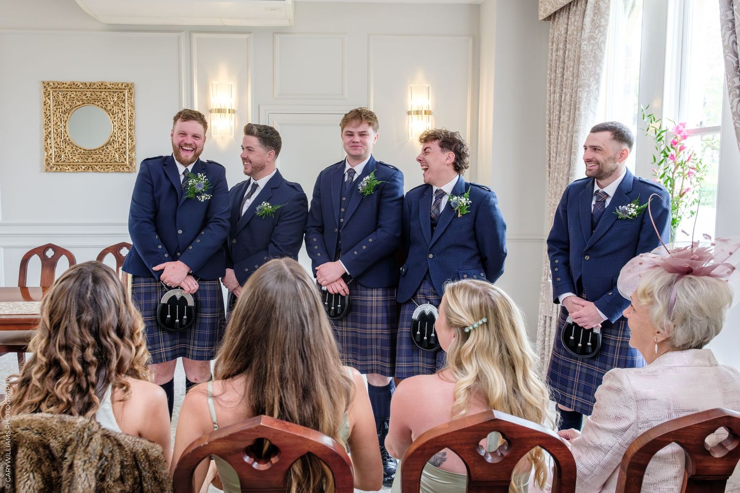 Five groomsmen in navy suits and kilts smiling at a wedding ceremony indoors.