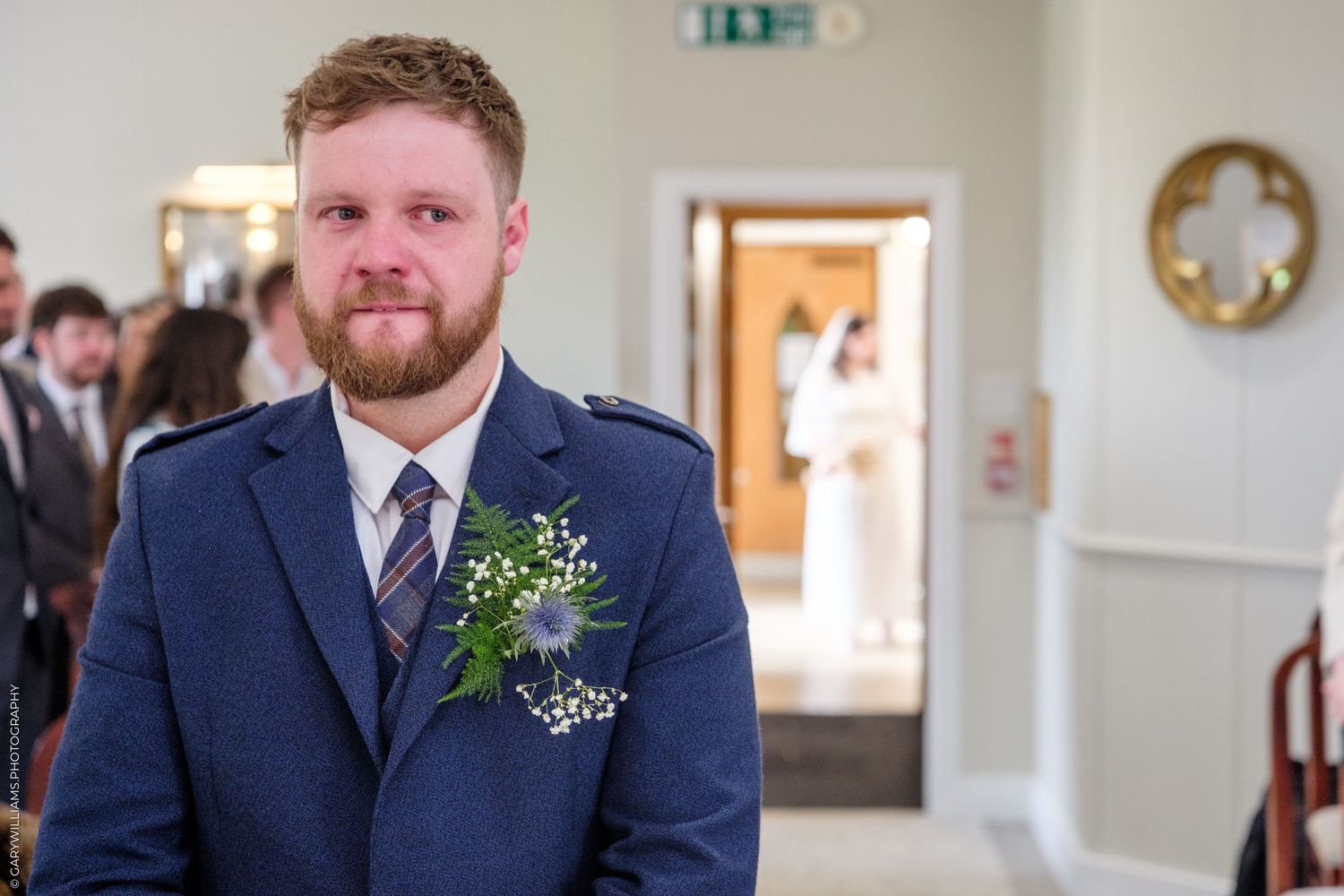 Groom in navy suit with floral boutonniere watches bride approach during wedding ceremony indoors.