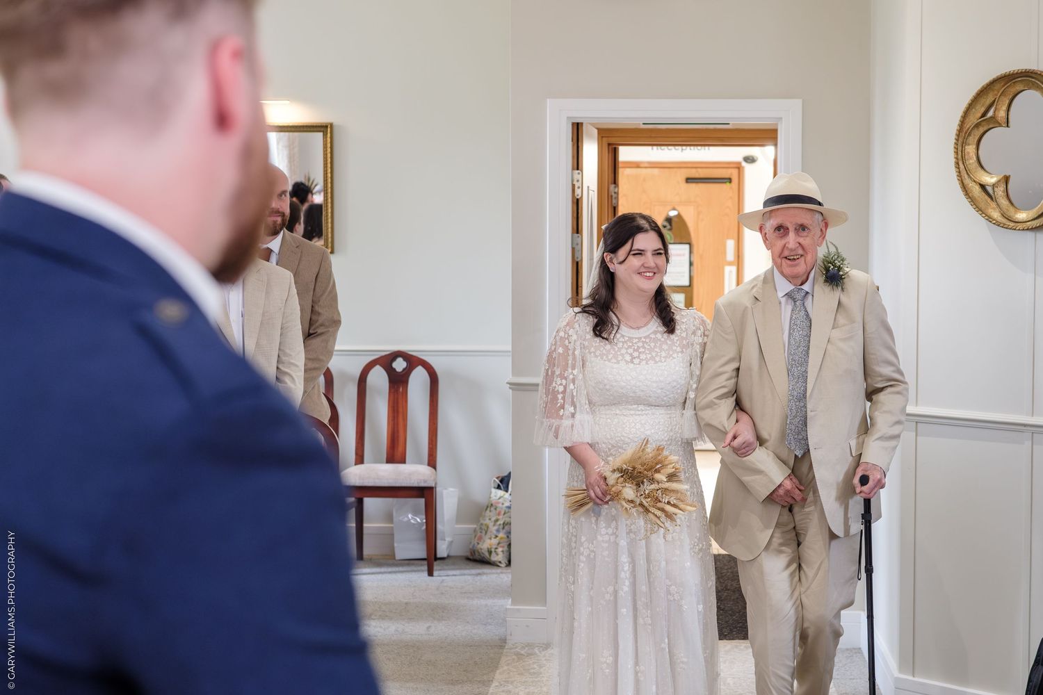 Bride in lace wedding dress walks down aisle with elderly man using cane during indoor wedding ceremony.