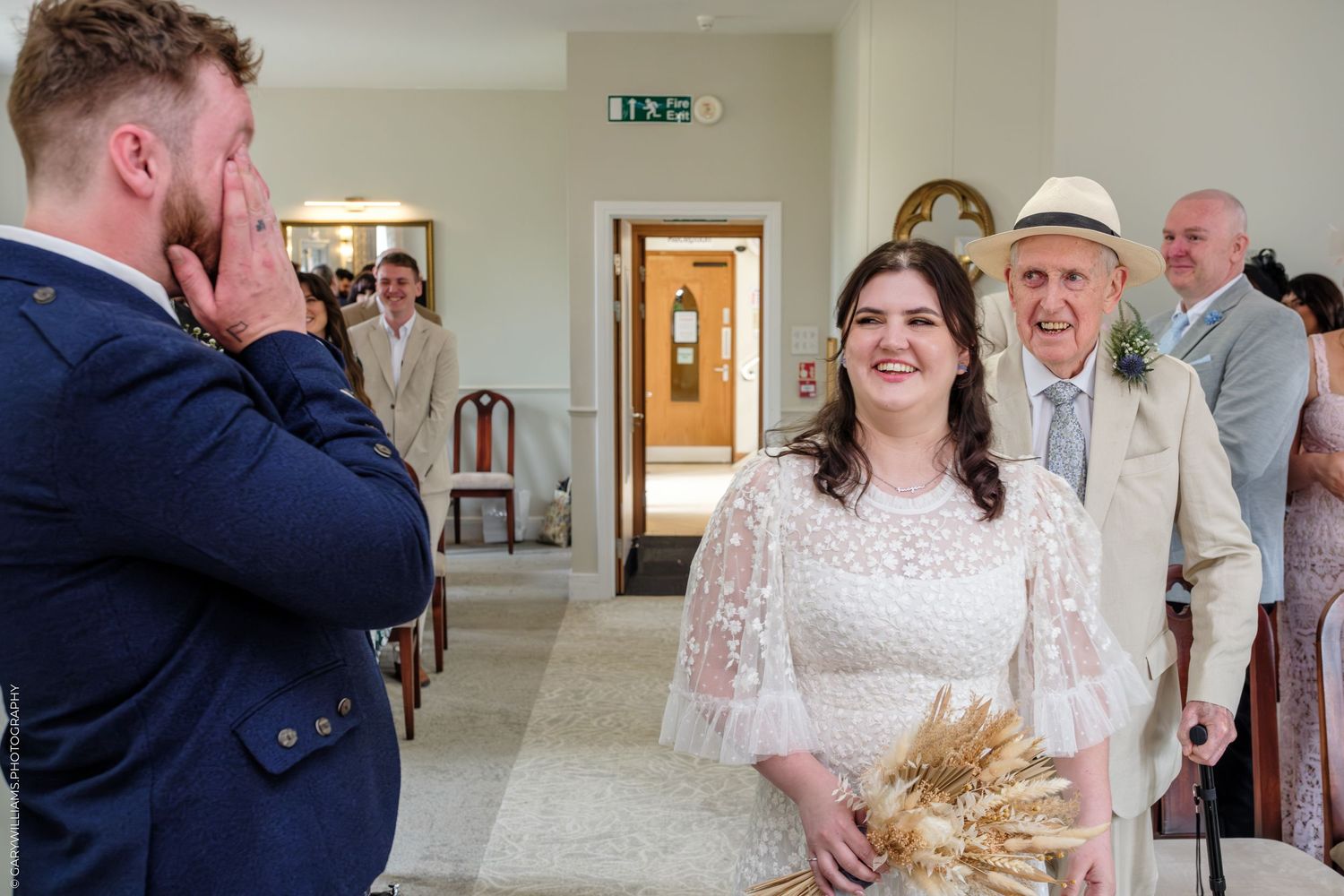 Emotional groom reacts as bride walks down aisle in white lace dress holding wheat bouquet at elegant wedding ceremony.