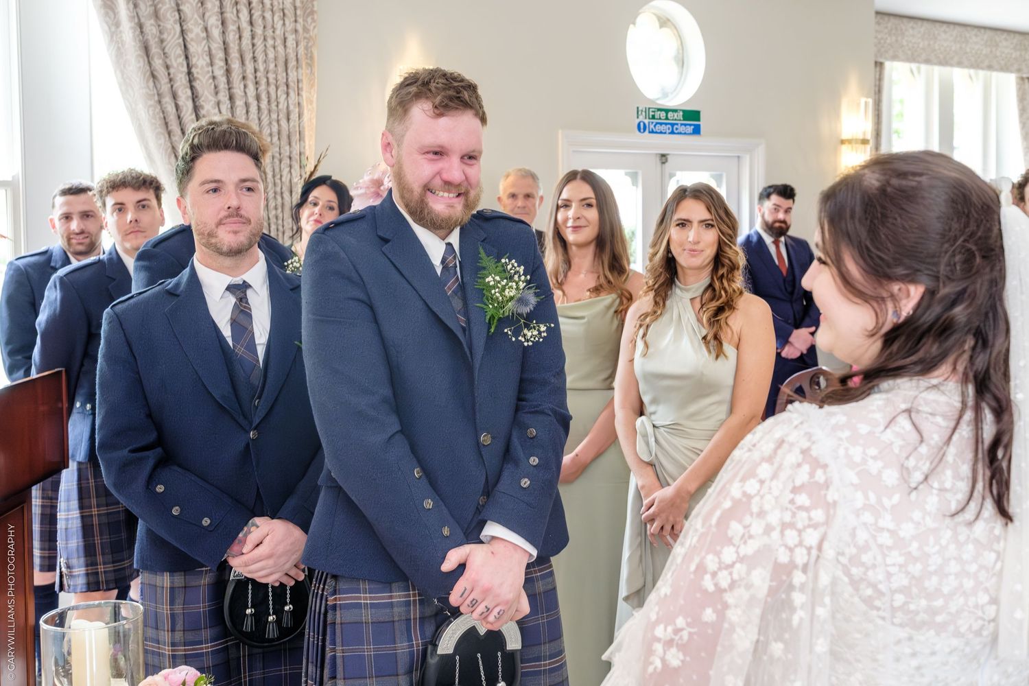 Groom in Scottish tartan kilt smiling at bride during wedding ceremony with guests in background.