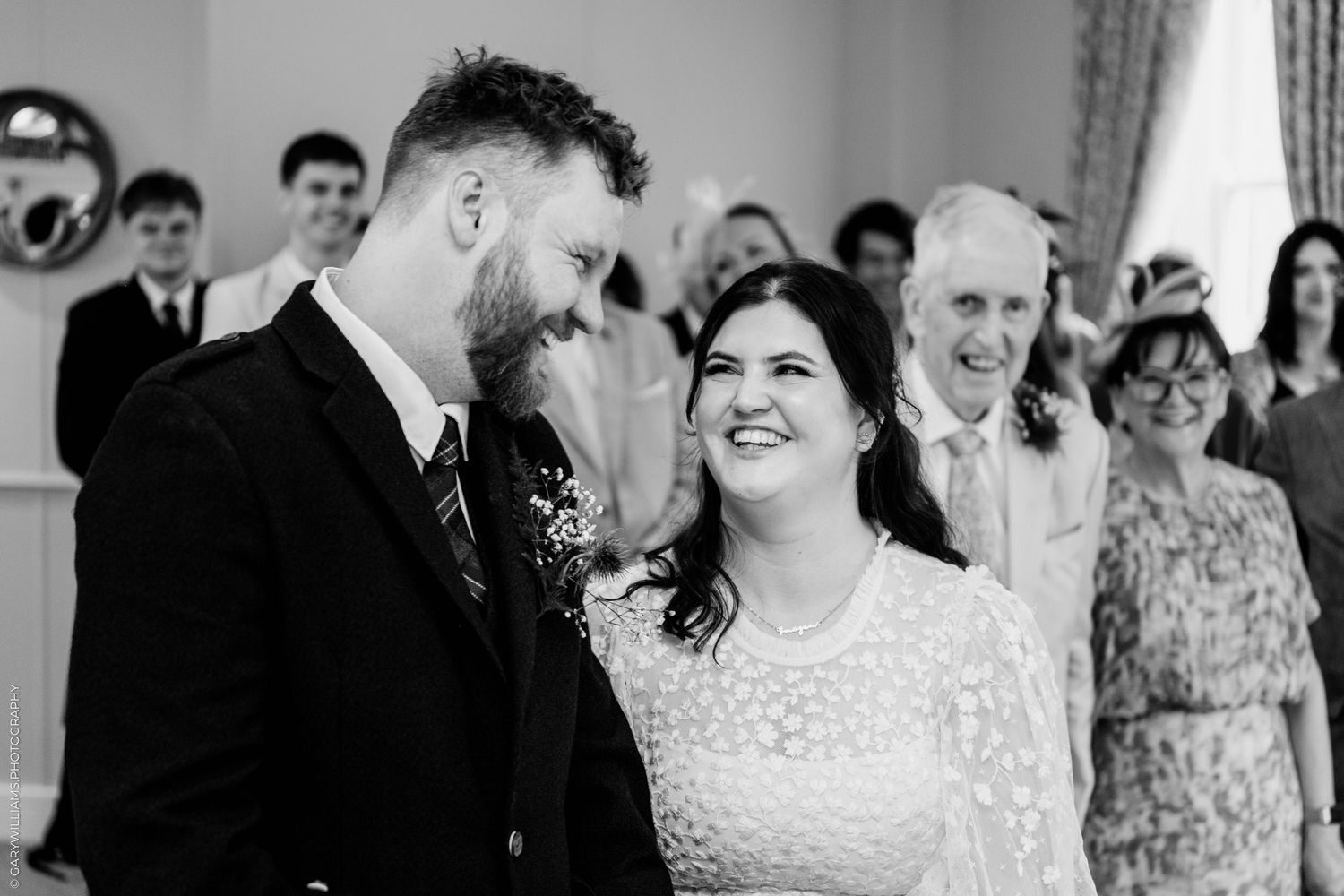 Black and white photo of a smiling bride and groom during their wedding ceremony with guests in the background.